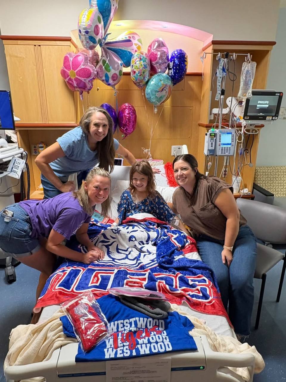 Grace in her hospital bed under a Westwood Tigers blanket, surrounded by balloons, with three women gathered around her.