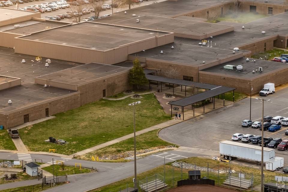 Aerial view of a school campus with tan brick buildings, rooftop vents, a covered outdoor walkway, and parking lots on either side.