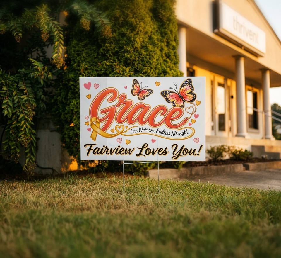 A Grace, Fairview Loves You! yard sign with orange lettering and butterflies stands in the grass outside a Fairview business.