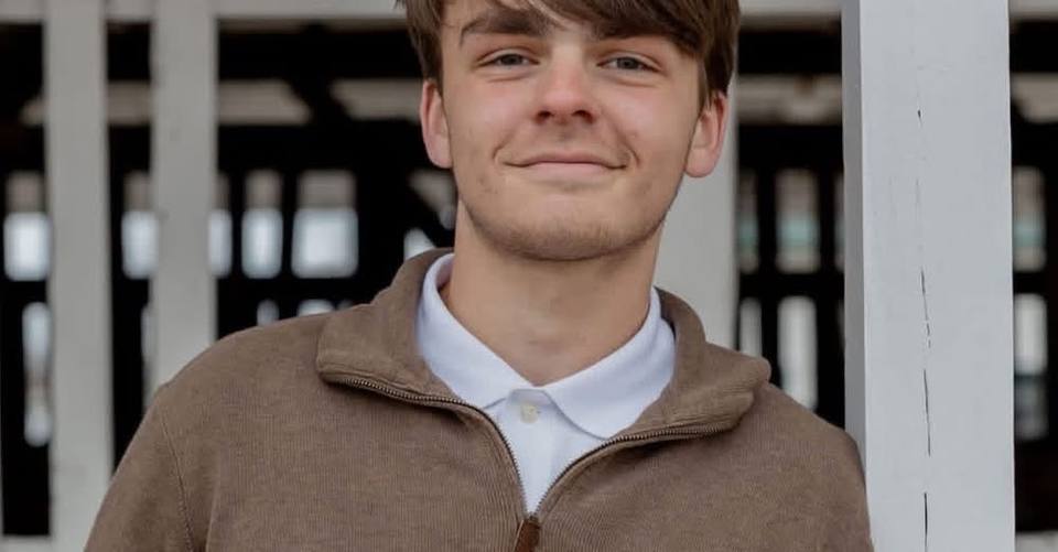 A smiling young man in a brown zip sweater over a white collared shirt, standing on a porch.