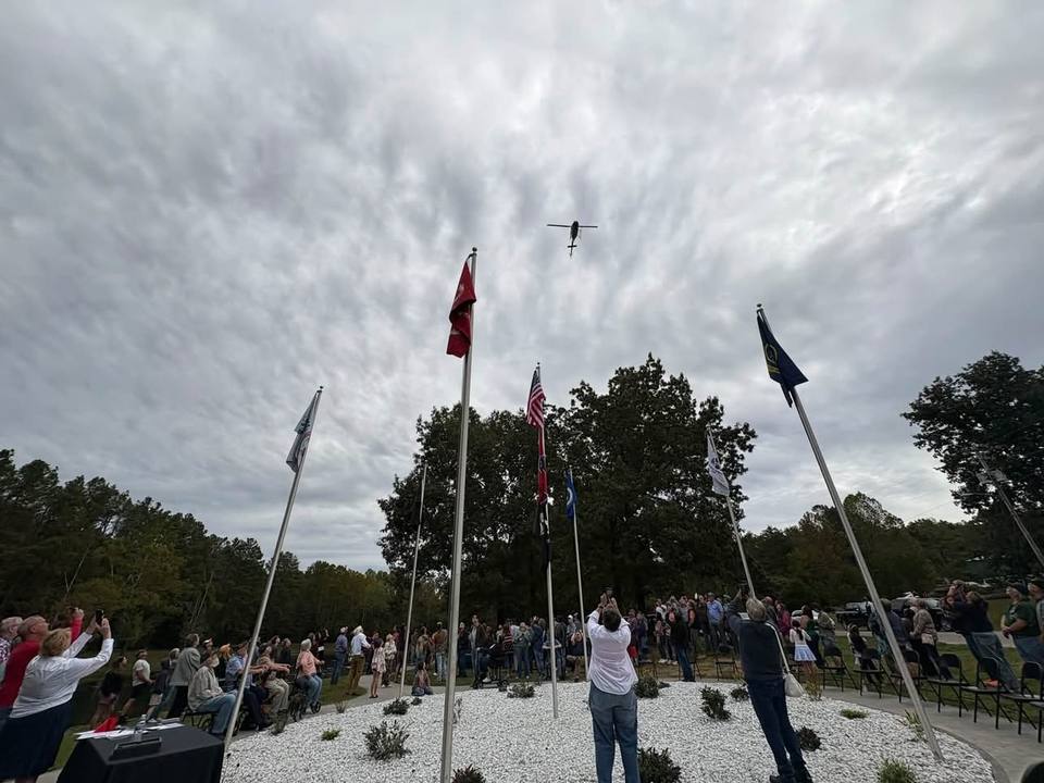 A crowd gathers around flagpoles at Veterans Memorial Park, watching a helicopter fly overhead during the grand opening ceremony.