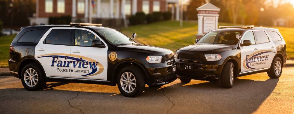 Two Fairview Police Department vehicles parked outside city hall in low evening sun.