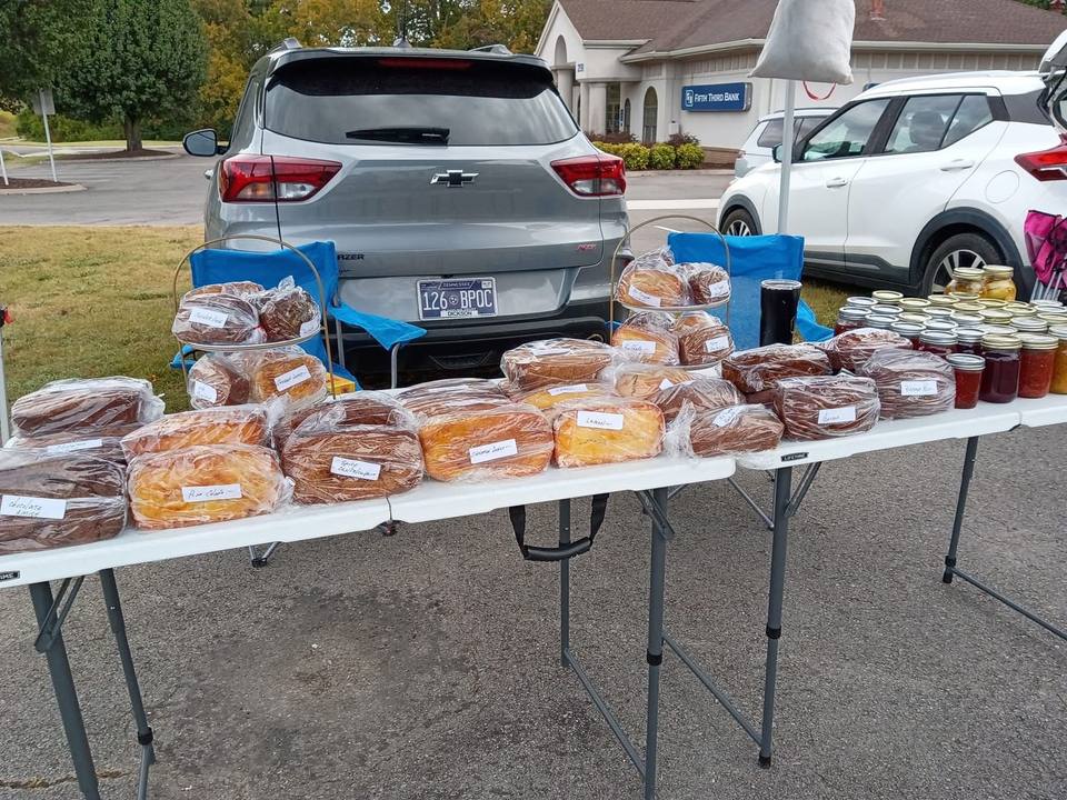 A market table lined with loaves of bread and jars of jam and preserves, with cars parked behind it.