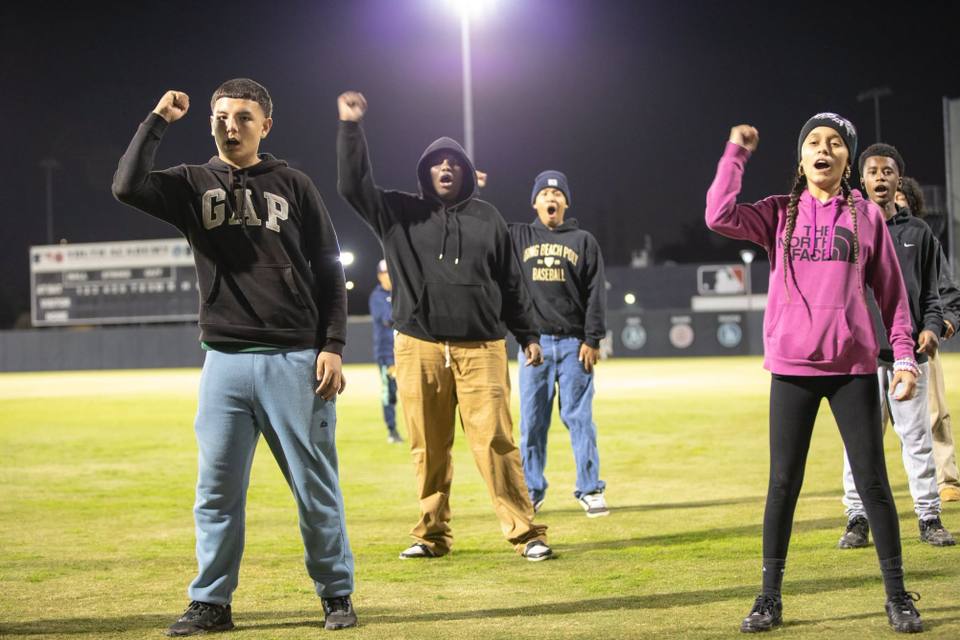 Several teenagers in hoodies stand on a baseball field at night under stadium lights, each raising a fist and shouting, with a scoreboard behind them.