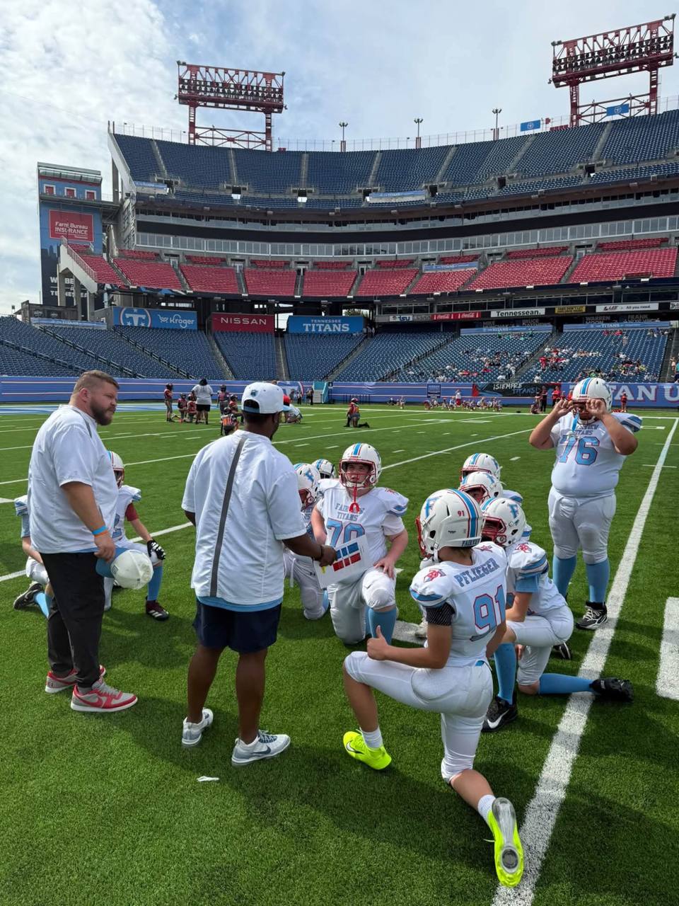 Football players in white and light blue Fairview Titans uniforms kneel on the turf around two coaches at Nissan Stadium, with mostly empty stands behind them.