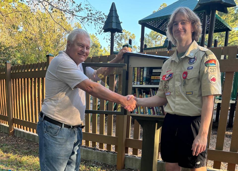 Friends of the Fairview Library President Gerry Suchomski shakes hands with Eagle Scout Jackson Jones beside a Little Free Library full of books.