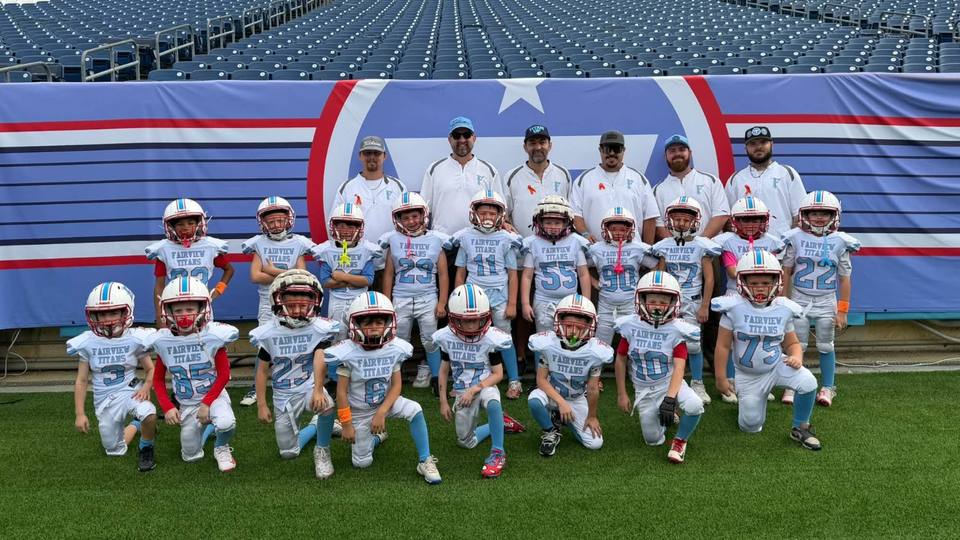 Fairview Titans youth football players in white and light blue uniforms kneel and stand in two rows in front of a Tennessee Titans banner at Nissan Stadium, with six coaches standing behind them.