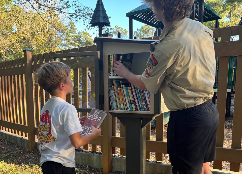 A young reader holds up a book picked out of a Little Free Library while Jackson Jones stocks its shelves.