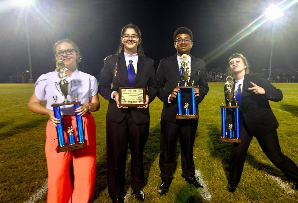 Four band students stand on a football field at night under stadium lights, three holding tall blue and gold trophies and one holding a plaque.