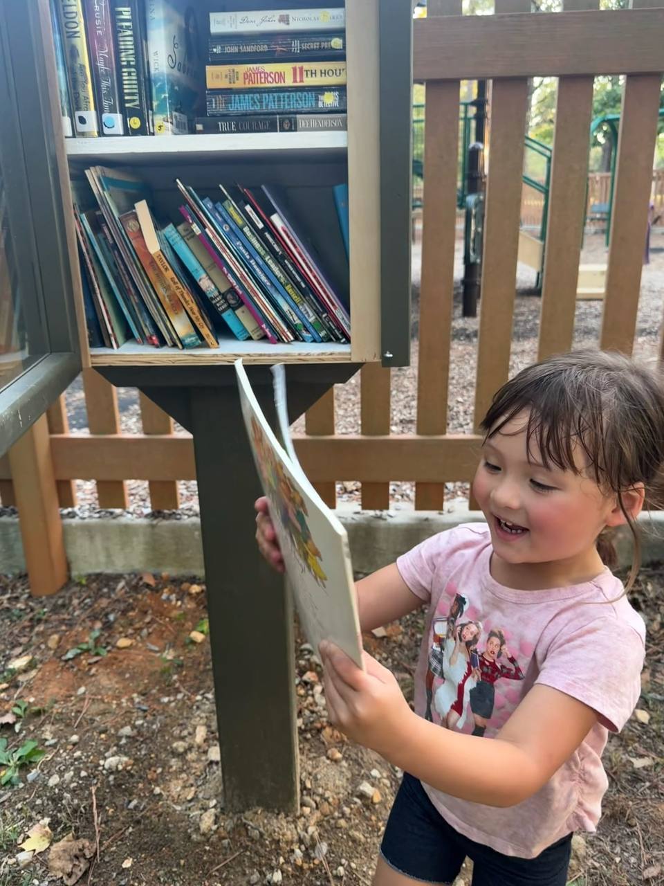 A smiling young girl pulls a picture book out of a Little Free Library stocked with books.